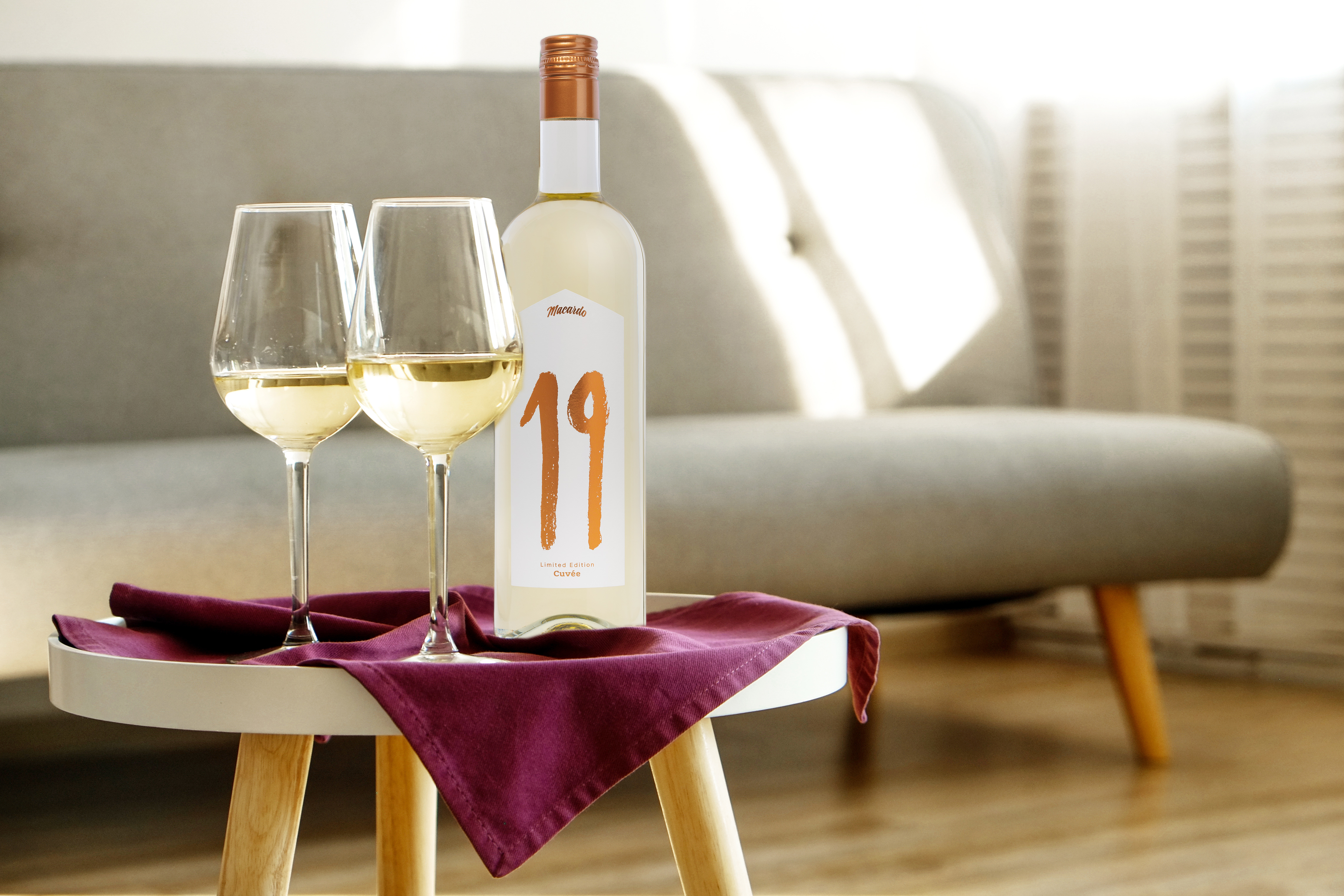 Zwei Gläser Weisswein und eine Flasche "19" Cuvée auf einem Tisch mit violettem Tuch. Die Flasche mit goldener Schriftzug "19" im Fokus, der helle Wein schimmert in den Gläsern. Im Hintergrund ein Sofa. Einladende Szene für Genuss.